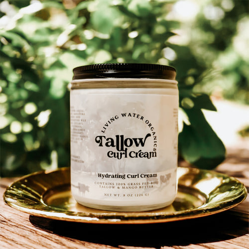 Tallow Curl Cream in glass jar with metal tin lid sitting on a gold plate outside on a wooden table with green leaves behind it.